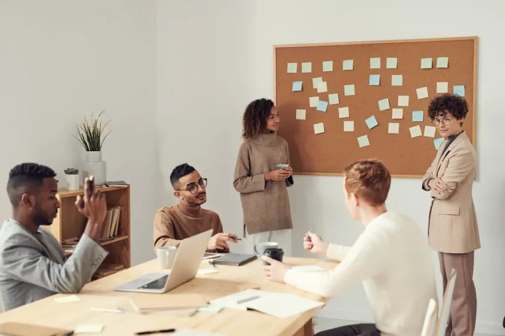 Set Clear Objectives A diverse group of colleagues having a meeting in a modern office, with sticky notes on a corkboard and laptops on the table.